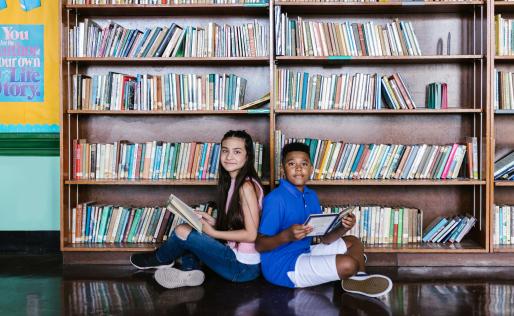 Een jongen en een meisje zitten rug aan rug voor een boekenkast