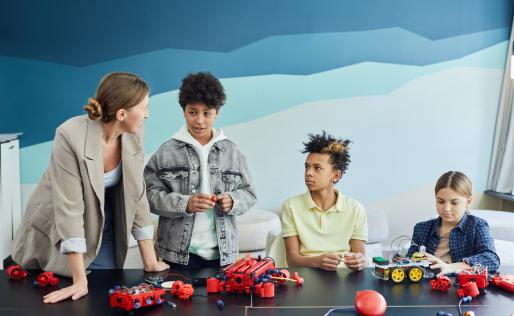 Drie kinderen en een juffrouw zitten aan tafel en zijn bezig met draden.
